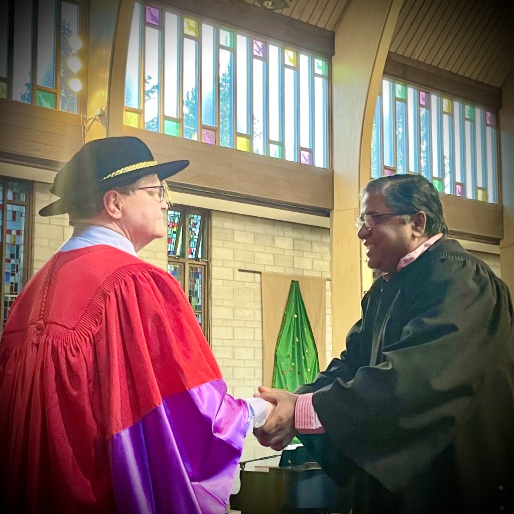 Graduation photo of a gowned graduate smiling and enthusiastically shaking hands with a gowned academic official