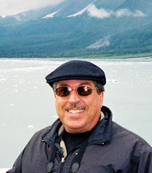 A smiling man wearing mirrored sunglasses standing outdoors in front of a bay and a mountain