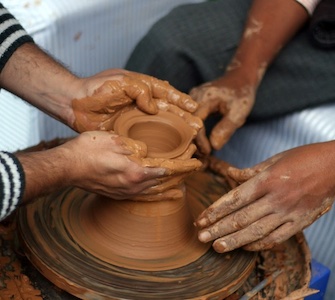 Two pairs of hands working together at a potter's wheel shaping clay illustrating a post on the economics of creation
